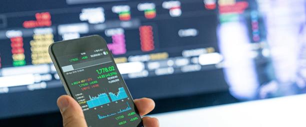 Pessoa segurando um smartphone que exibe dados do mercado de ações em frente a uma tela de negociação