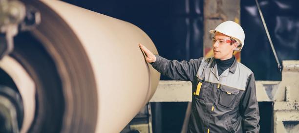 Worker in safety gear inspecting large roll of paper in a production facility.