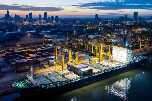 Porto de contêineres à noite com guindastes, navios de carga e o horizonte da cidade ao fundo.
