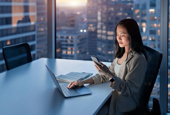 Professional Asian businesswoman using smartphone and laptop at office during evening, with city skyline in background.