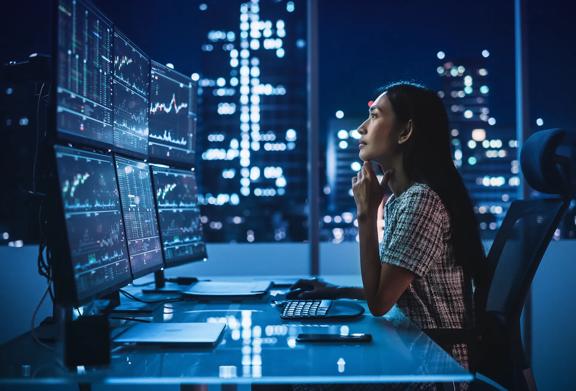 Woman analyzing financial data on multiple monitors at night, with city lights in the background