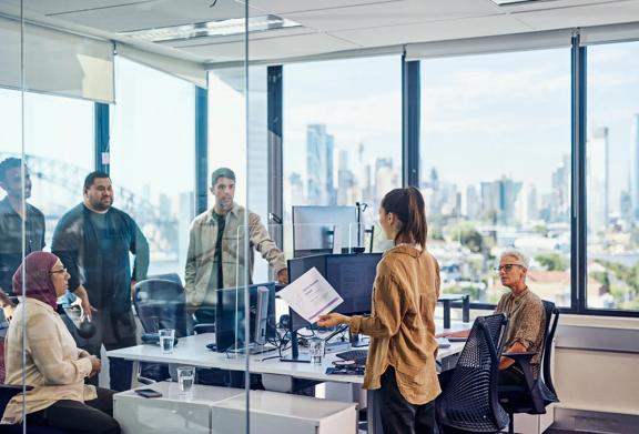 Woman standing and holding a document while presenting to a group of colleagues in a glass-walled office with city skyline view