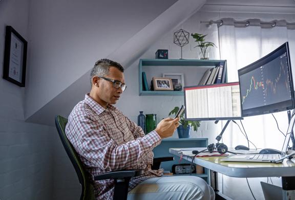 Man working from home analyzing financial charts on dual monitors while using a smartphone