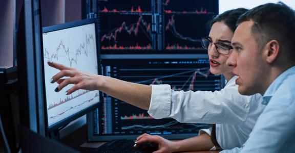 Two financial analysts reviewing and discussing stock market data on multiple computer monitors