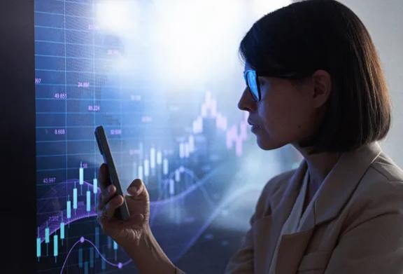 Mulher com óculos analisando gráfico do mercado de ações em grande tela digital usando smartphone