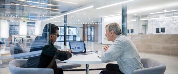 Two business professionals sitting at a round table in a bright, modern office space reviewing graphs and financial charts on a laptop screen through a glass partition.