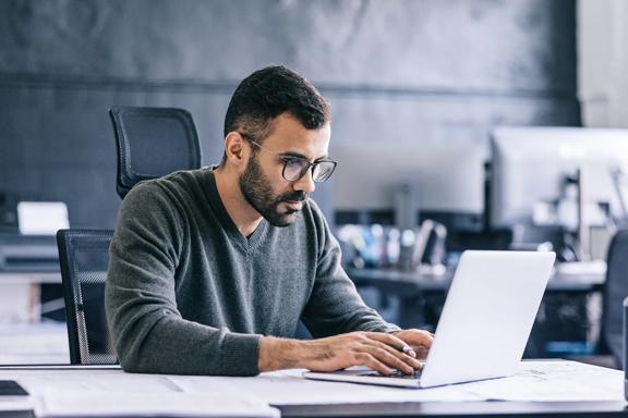 Homem jovem com óculos, sentado em escritório moderno, digitando no laptop com foco em suas tarefas de trabalho.