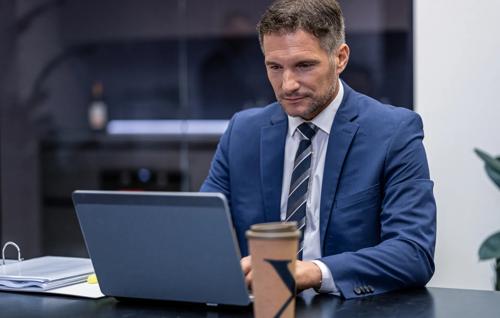 Focused businessman in a suit working on a laptop at a modern office desk with coffee cup nearby.