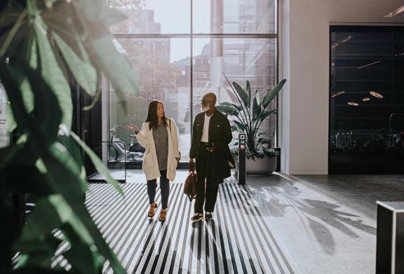 Two professionals walking and talking in modern office lobby with natural light and large indoor plants.