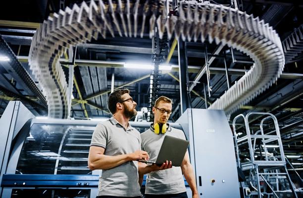 Engineers analyzing production data on a laptop in an automated industrial facility with overhead conveyor system.
