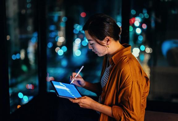 Person using a digital tablet with a stylus in front of a large window overlooking a cityscape at night. The tablet screen displays a chart or graph.