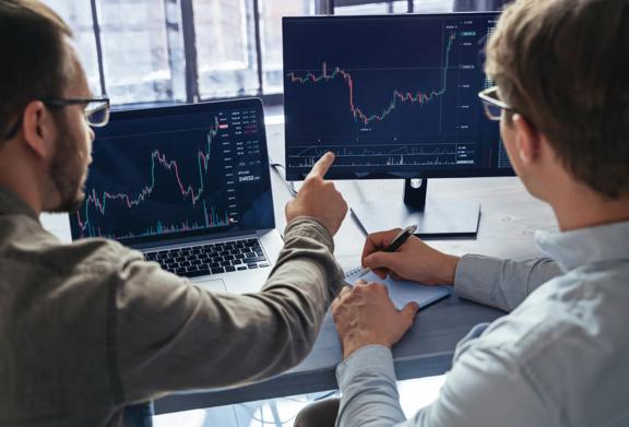 Dois profissionais analisando gráficos financeiros em várias telas em um escritório moderno