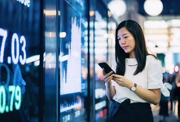 Uma mulher analisa dados financeiros em um smartphone enquanto está em frente a uma tela digital do mercado de ações