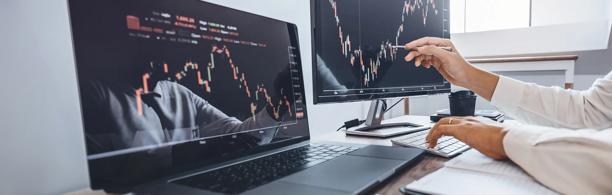 Two traders analyzing candlestick stock charts on dual monitors in a modern office setting, reviewing market trends and financial data.
