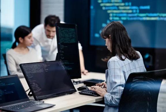 Mulher programando em múltiplas telas de computador enquanto colegas conversam ao fundo