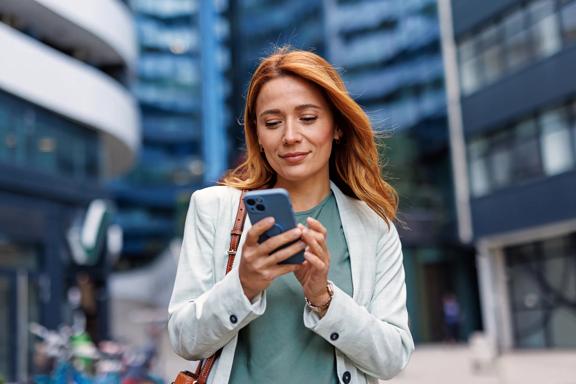 Mulher ruiva de negócios caminhando em área urbana moderna, sorrindo enquanto usa smartphone para comunicação.
