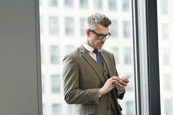 Homem executivo de terno e óculos, em escritório corporativo, utilizando smartphone próximo à janela.