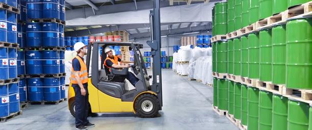 Warehouse workers operating a forklift to transport barrels of green and blue chemicals, ensuring safe handling in an industrial setting.