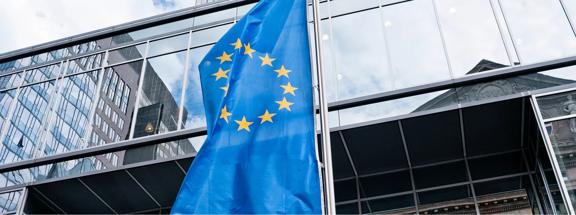 European Union flag waving in front of a modern glass office building in an urban setting.