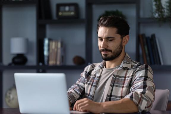 Homem jovem sentado em home office, usando laptop para trabalho remoto, com estante e livros ao fundo.