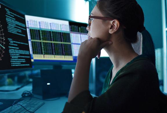 Person working on dual monitors displaying financial data and stock market charts in a modern office.