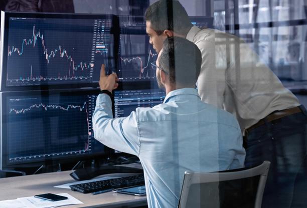 Dois profissionais financeiros analisando gráficos do mercado de ações em vários monitores em um escritório moderno