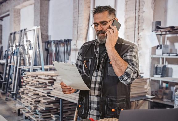 Smiling carpenter or craftsman talking on the phone while reviewing paperwork in a woodworking shop