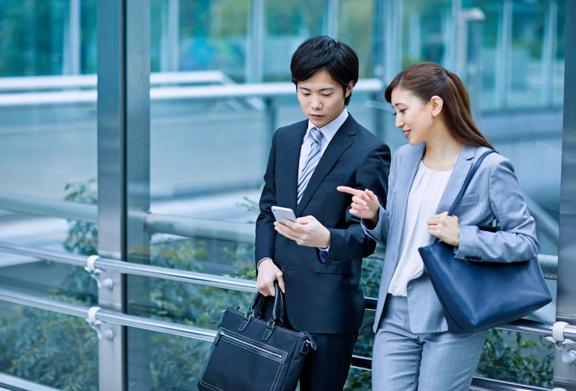 Two business professionals standing in a modern glass building, reviewing information on a smartphone while carrying briefcases and handbags.