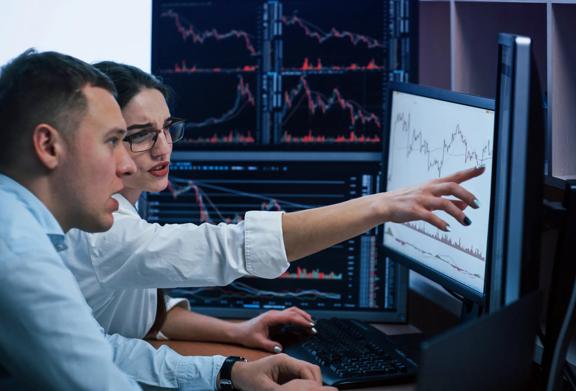 Two financial analysts examining stock market charts on multiple computer monitors