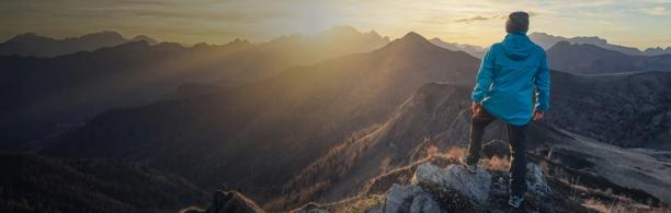 Homem em pé no cume de uma montanha ao nascer do sol, com vista para uma dramática paisagem alpina e picos iluminados pelo sol.