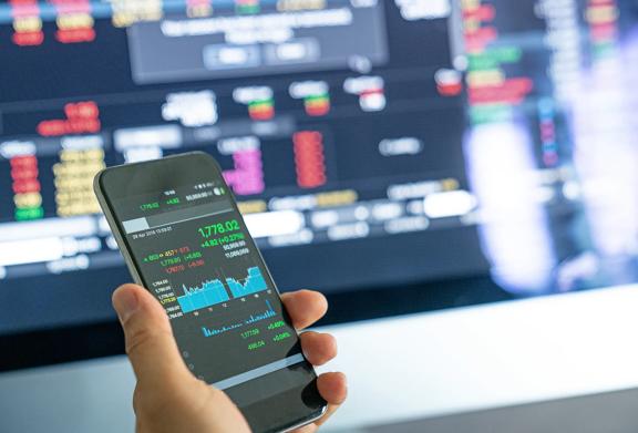 Person holding a smartphone displaying stock market data in front of a trading screen