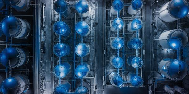 Aerial view of a large industrial facility with multiple blue storage tanks, interconnected pipelines, and processing infrastructure, representing energy production, commodity storage, industrial operations, and supply chain logistics.