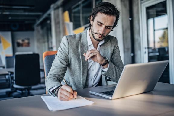 Profissional trabalhando em uma mesa, fazendo anotações em papel enquanto utiliza um laptop em um ambiente de escritório moderno.