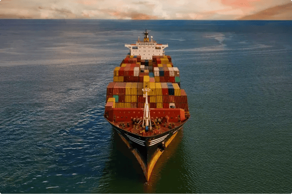Front-facing aerial view of a large container ship carrying colorful shipping containers across calm ocean water.