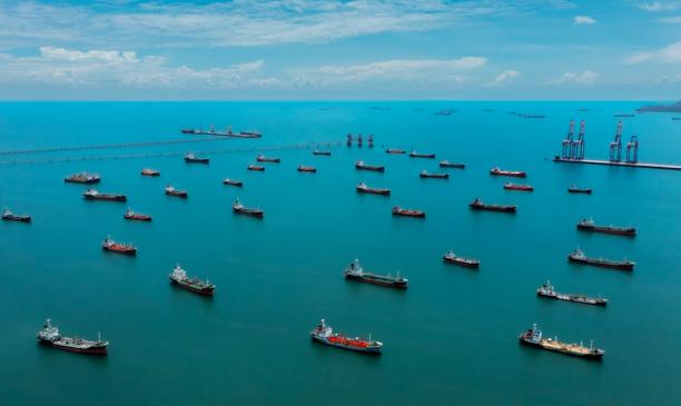 Aerial view of numerous cargo ships and vessels anchored across a wide turquoise sea near the coastline.