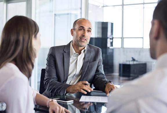Consultor de negócios reunido com dois clientes em um escritório com paredes de vidro, discutindo estratégias e planejamento financeiro.