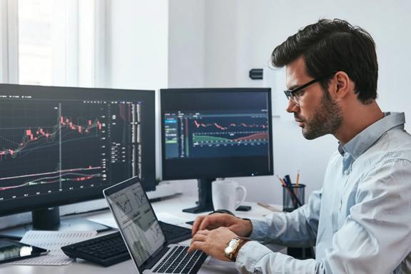 Professional trader working at laptop with 2 desktop screens showing financial charts