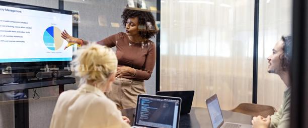 A diverse group of professionals in a glass-walled office meeting room engaged in a data-driven presentation, with a woman presenting a pie chart on a screen while two colleagues with laptops attentively listen.