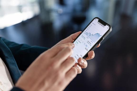 Close-up de mãos segurando smartphone com gráfico financeiro exibido na tela, representando análise de mercado de ações.