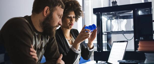 Two professionals discussing a 3D-printed object in a modern office, with a 3D printer visible in the background