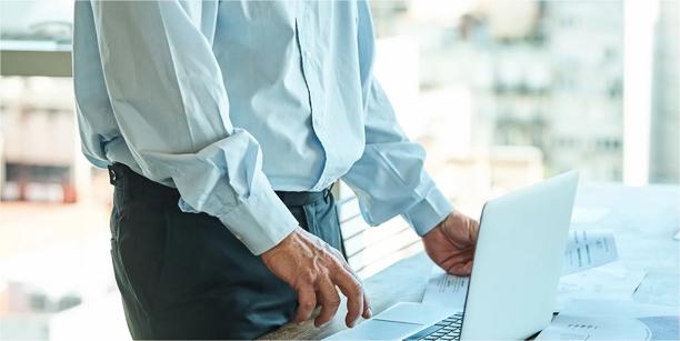 Person standing at a desk with one hand on a laptop, wearing a light shirt in a bright office setting.