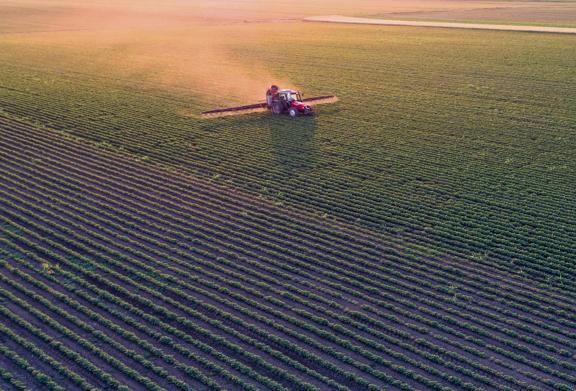 Aerial view of a tractor spraying crops in a large agricultural field at sunset