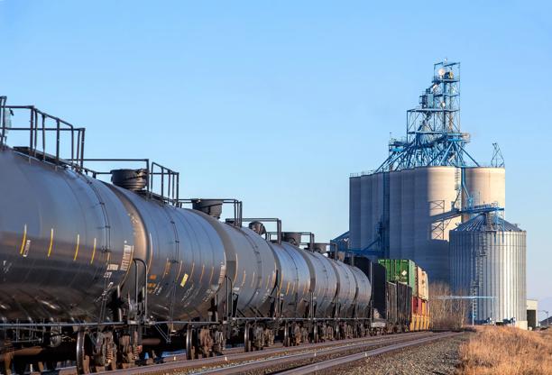 Vagões-tanque pretos em trilhos de trem ao lado de grandes silos de grãos e instalações de processamento agrícola