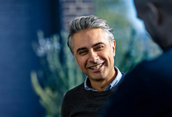 Confident middle-aged man with gray hair smiling warmly while speaking to a colleague in a casual yet professional setting, with soft natural lighting in the background.