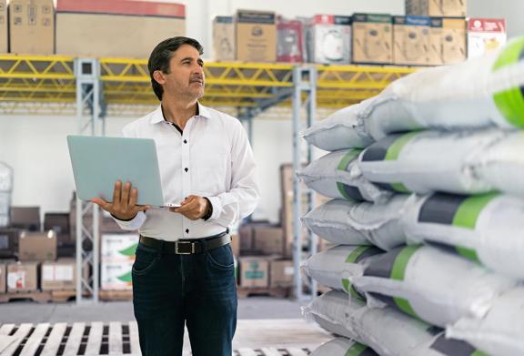 Warehouse manager using a laptop to manage inventory, standing beside stacked bags of goods in a storage facility