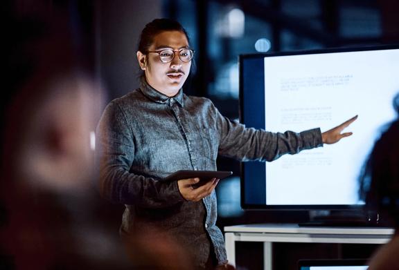 Man wearing glasses giving a presentation with tablet in hand while pointing to screen with projected slides in office