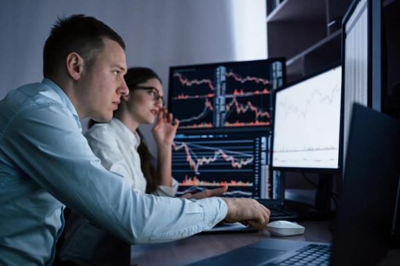 Two traders analysing financial charts and market data on multiple monitors in a trading office