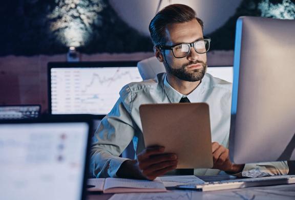 Business professional wearing glasses analyzing financial charts and data on a desktop monitor while holding a tablet, working late in a tech-driven office environment.