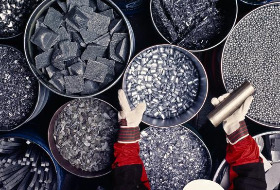 Worker handling various forms of refined metal including pellets, shavings, and bars in industrial containers