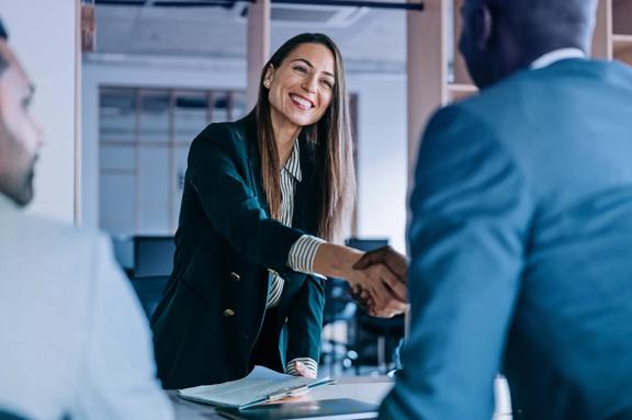 A female professional in an office setting, shaking hands with a male professional across a desk 